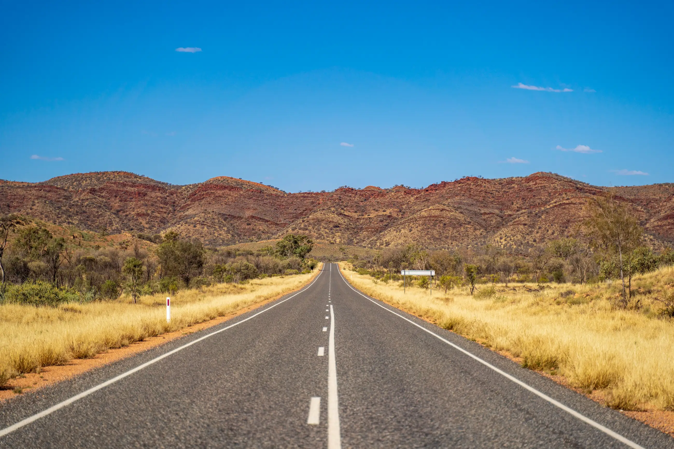 A sealed outback highway running straight toward red rock ranges under a clear blue sky